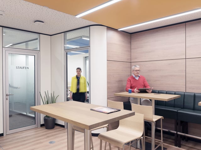 Raum mit Stehtischen, Sitzbank an einer Holzwand und Glastür mit der Aufschrift „Staufen“, zwei Personen