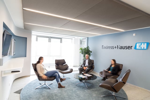 Wartebereich mit braunen Ledersesseln auf einem runden Teppich vor einer Wand mit dem Schriftzug Endress+Hauser, drei Personen sitzen