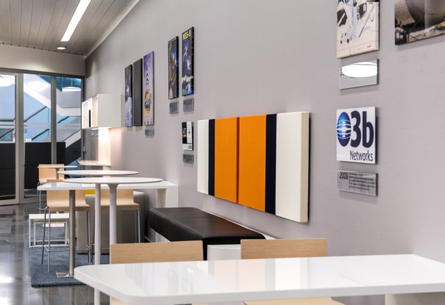 Weiße Stehtische und Sitzbank vor einer Wand mit Bildern, einem orange-blauen Wandpaneel und dem Logo „O3b Networks“