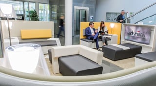 Lounge im Foyer mit weiß-grauen Sitzmöbeln, gelben Rückenlehnen und Stehleuchten, zwei Personen im Gespräch, rechts ein Bildschirm mit dem Logo adwerk