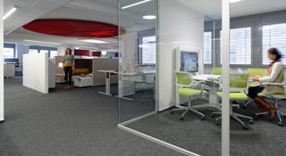 Offene Bürofläche mit grauem Teppich, einem roten runden Deckensegel und einem Besprechungsraum hinter Glas mit hellgrünen Stühlen