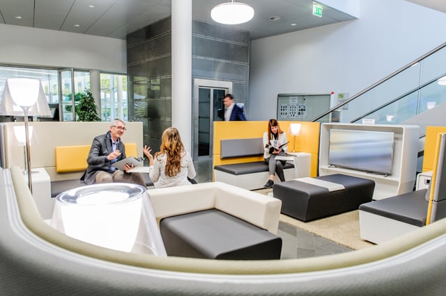 Lounge mit weißen und gelben Sitzmöbeln, Besucher im Gespräch, hinten eine Treppe