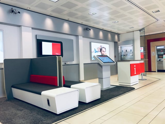 Kundenhalle mit Sitzbank, Terminal mit Touchscreen und roter Infotheke mit „i“-Symbol