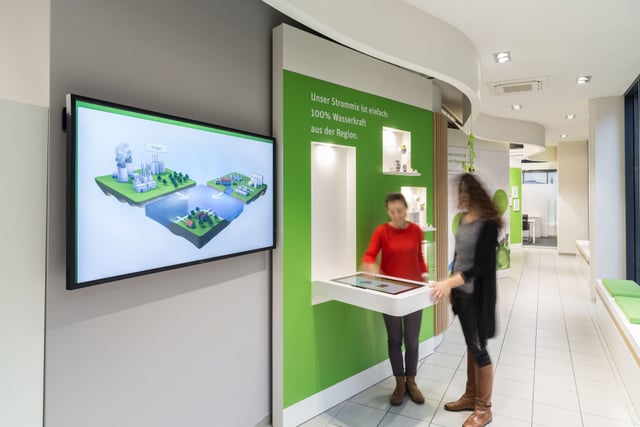 Zwei Frauen an einem Tisch mit Touchscreen vor einer grünen Wand, daneben ein Bildschirm