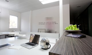 Helles Büro mit weißen Schreibtischen, Laptop und Rollkartei, rechts ein Sideboard mit einer Schale grüner Äpfel und einem Tablet. An der Wand der Schriftzug: Qualität ist, wenn die Kunden zurückkommen und nicht die Ware.