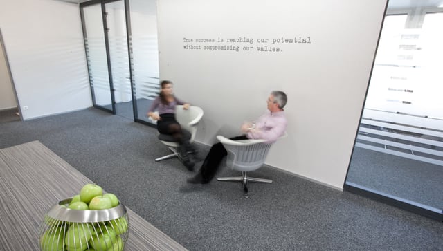 Zwei Personen auf Drehsesseln vor einer Wand mit dem Spruch „True success is reaching our potential without compromising our values.“, vorne eine Schale Äpfel