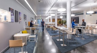 Langgestreckte Cafeteria mit Stehtischen, Barhockern, einem Sofa und blauen Teppichen, an der Wand Bilder und eine Vitrine, mehrere Personen im Gespräch