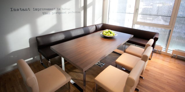 Besprechungsecke mit Holztisch, Eckbank und hellbraunen Stühlen, an der Wand der Spruch „Instant improvement is better than postponed perfection.“