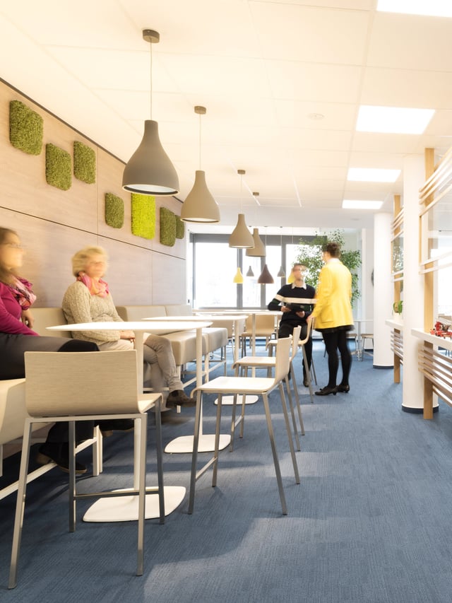 Café-Bereich mit Sitzbank, Stehtischen, Moosbildern an der Holzwand und Personen im Gespräch