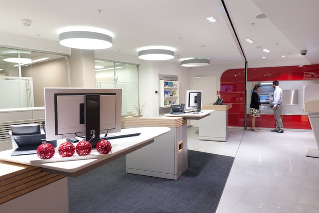 Kundenhalle mit Beratungstisch und Bildschirm, im Hintergrund Personen an Automaten