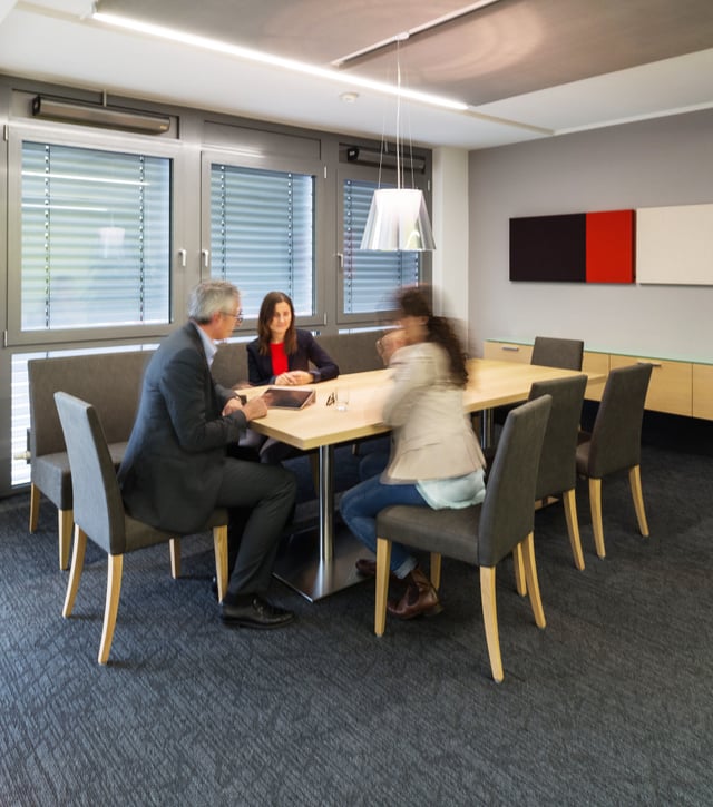 Drei Personen im Gespräch an einem Holztisch mit grauen Polsterstühlen