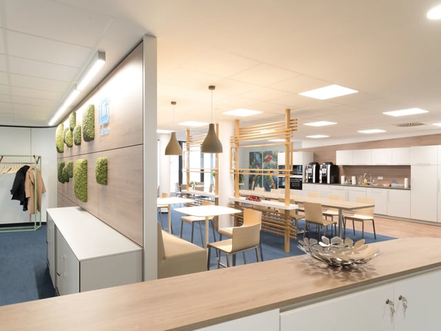 Blick über eine Theke in einen Café-Bereich mit Küchenzeile, Tischen und Moosbildern